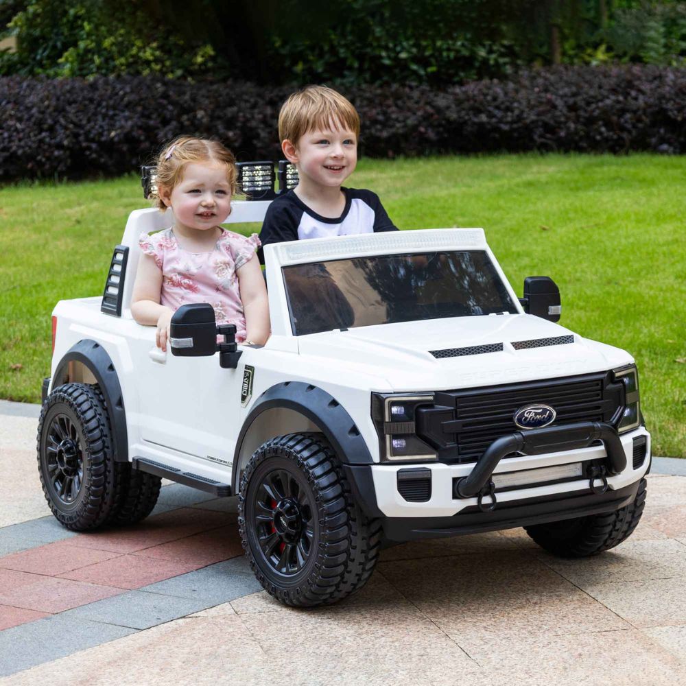 Two children sitting in a white toy truck outdoors on a paved area with grass and bushes in the background.